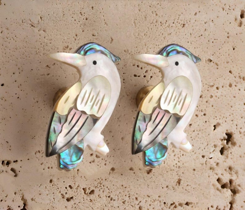 May include: A pair of bird-shaped cabinet knobs. Each knob has a white body with iridescent abalone shell accents on the wings and tail. The knobs have a gold-coloured base and a blue iridescent crest.