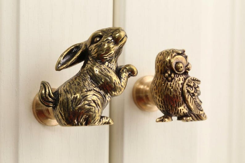 May include: Two bronze cabinet knobs, one shaped like a rabbit and the other like an owl. The rabbit is in a playful pose, and the owl has detailed feathering. Both knobs have a brass finish and are mounted on a white surface.