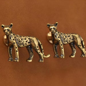Pode incluir: Dois puxadores de armário em forma de leopardo em latão. Os puxadores são detalhados com manchas e são montados em uma base redonda. O fundo é de cor marrom sólida.