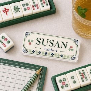 May include: A rectangular acrylic place card featuring the name SUSAN and the text Table 4 in dark green font. The design includes a decorative border with green and blue diamond patterns, surrounded by Mahjong tiles and a green striped pen.