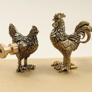 May include: Two bronze-colored rooster figurines. One is a hen with a bottle stopper attached. The other is a rooster with detailed feathering and a prominent comb. Both are standing on a light brown surface.