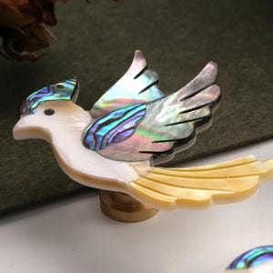 May include: A decorative bird figurine with iridescent wings and tail feathers. The bird's body is white, and the wings and tail feature a shimmering, multi-coloured abalone shell design. The figurine is mounted on a small wooden base.