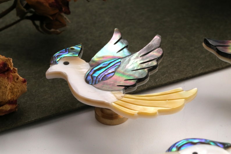 May include: A decorative bird figurine with iridescent wings and tail feathers. The bird's body is white, and the wings and tail feature a shimmering, multi-coloured abalone shell design. The figurine is mounted on a small wooden base.
