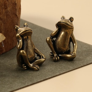 Könnte beinhalten: Zwei Froschfiguren aus Bronze in entspannter Pose, eine mit der Hand am Kinn. Die Frösche sitzen auf einer grauen Oberfläche, mit einem Holzblock und Blättern im Hintergrund. Die Frösche haben ein metallisches Finish.