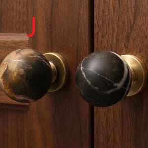 Op de afbeelding: Twee bolvormige kastknoppen van donkerbruine en zwarte steen met witte aders, gemonteerd op ronde voetjes van geborsteld messing. De knoppen zijn bevestigd aan een houten kastdeur, met een rode letter J bij de linkerknop.