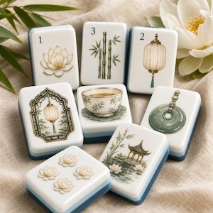 May include: A set of white rectangular mahjong tiles featuring detailed illustrations of lotus flowers, bamboo stalks, lanterns, a tea cup, a jade ring, and a pavilion. The tiles have blue-tinted bases and are arranged on a textured fabric surface.