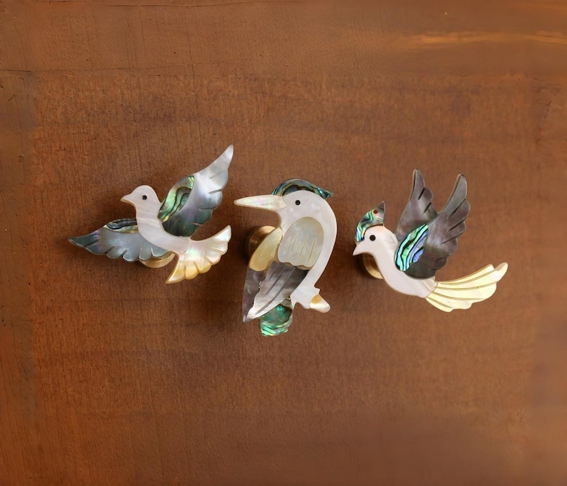 May include: Three decorative bird-shaped cabinet knobs. Each knob features a unique design with iridescent shell accents in shades of green, blue, and gold. The birds are set against a brown background, adding a touch of nature-inspired elegance to any furniture.