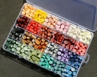 Peut inclure: Une boîte en plastique transparente remplie de 24 perles de cire de différentes couleurs. Les perles sont disposées en grille et sont toutes de la même taille et de la même forme. La boîte est ouverte et les perles sont visibles.