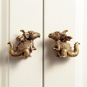 May include: Two brass-toned metal cabinet knobs shaped like small dragons. Each knob features a textured, scaly body, small wings, a curved tail, and a detailed head with prominent eyes, mounted on a vertical white panelled surface.