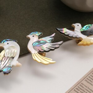 May include: Three decorative bird ornaments with iridescent shell detailing. The birds feature white, yellow, and black accents, with a pearlescent sheen. The ornaments are arranged on a white surface.