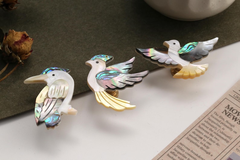 May include: Three decorative bird ornaments with iridescent shell detailing. The birds feature white, yellow, and black accents, with a pearlescent sheen. The ornaments are arranged on a white surface.