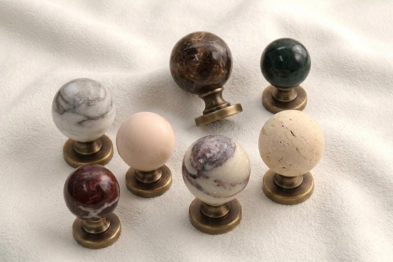 Op de afbeelding: Een verzameling van zeven bolvormige kastknoppen met diverse natuurstenen afwerkingen, waaronder wit marmer, donkerbruin, diepgroen, effen beige, rood marmer en een wit met paars gemarmerd patroon, allemaal gemonteerd op ronde messing voetjes.