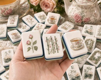May include: A set of rectangular mahjong tiles featuring white surfaces with various floral, botanical, and teacup illustrations in soft colors. Each tile has a layered base with blue and green bands. The tiles are arranged on a lace surface.