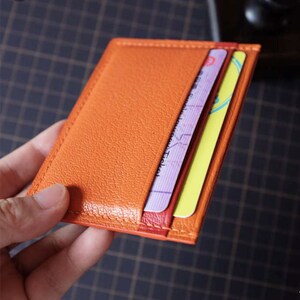 Op de afbeelding: Oranje leren kaarthouder met een gestikte rand. De houder heeft twee kaarten erin gestoken, een met de tekst "Ticket" en de andere met Chinese karakters.