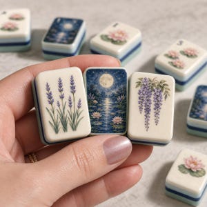 Può includere: Tre tessere rettangolari da mahjong con angoli arrotondati. La tessera a sinistra presenta steli di lavanda viola, quella centrale raffigura una luna piena su uno stagno di loto e quella a destra mostra fiori di glicine viola su fondo crema.