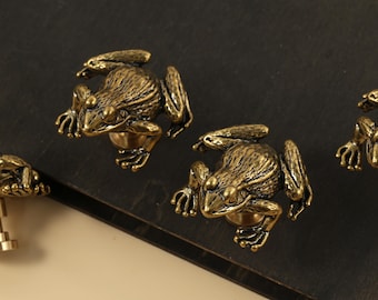 May include: Bronze frog cabinet knobs. The knobs are detailed with textured skin and realistic features. The frogs are in a crouching position, with their front legs extended. The knobs are mounted on a dark surface.