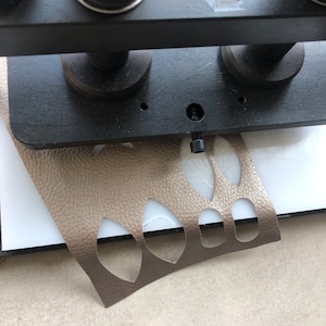 Op de afbeelding: Een close-up van een lederbewerkingstool met een stuk bruin leer met uitgesneden vormen die in de tool worden geperst.