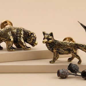 May include: Two brass animal-shaped cabinet knobs: a bear and a fox. The bear is on all fours, and the fox is standing. The knobs have a vintage, antique appearance. A few decorative elements are also present.