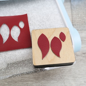 Op de afbeelding: Een close-up van een rode leren stansvorm met twee blaadjes en een kleine cirkel. De stansvorm ligt op een doorzichtig plastic vel met een glinsterend oppervlak. De stansvorm ligt naast een houten stansvorm met dezelfde uitgesneden vormen.