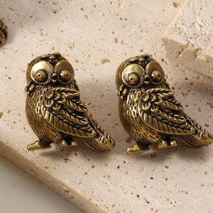 May include: Two antique gold-colored owl figurines. The owls have large, round eyes and detailed feather patterns. They are positioned on a light-colored, textured surface, suggesting a decorative or collectible item.