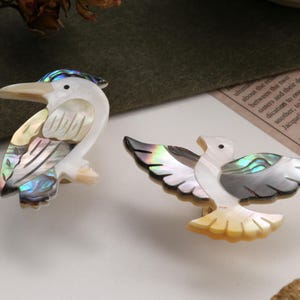 May include: Two decorative bird ornaments. One is a white and iridescent bird with a long beak. The other is a white and iridescent bird with outstretched wings. Both are made of a pearlescent material.
