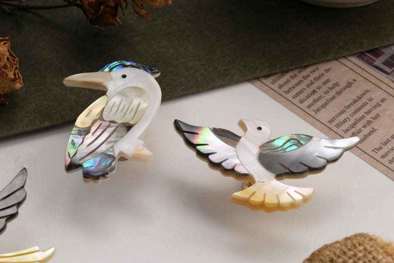 May include: Two decorative bird ornaments. One is a white and iridescent bird with a long beak. The other is a white and iridescent bird with outstretched wings. Both are made of a pearlescent material.