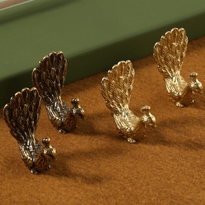 Könnte beinhalten: Vier goldene Pfauenfiguren mit detaillierten Federdesigns. Die Pfauen sind in einer Reihe auf einer braunen Oberfläche angeordnet.