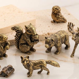 May include: A collection of small, antique brass animal figurines, including a rabbit, squirrel, owl, bear, moose, and fox. The detailed sculptures are displayed on a light-colored surface, showcasing their intricate designs and golden hue.