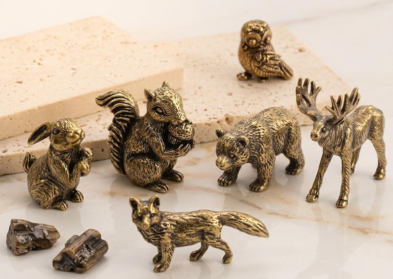 May include: A collection of small, antique brass animal figurines, including a rabbit, squirrel, owl, bear, moose, and fox. The detailed sculptures are displayed on a light-colored surface, showcasing their intricate designs and golden hue.