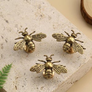 May include: Three antique brass bee-shaped cabinet knobs. Each knob features detailed wings and a round base for mounting. The knobs are arranged on a light-colored, textured surface, suggesting a decorative hardware theme.
