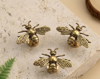 Könnte beinhalten: Drei antike Messing-Schrankknöpfe in Bienenform. Jeder Knopf hat detaillierte Flügel und eine runde Basis zur Montage. Die Knöpfe sind auf einer hellfarbigen, strukturierten Oberfläche angeordnet, was auf ein dekoratives Hardware-Thema hindeutet.