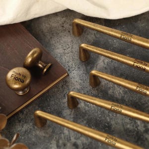 Op de afbeelding: Antieke messing kastbeslagset, inclusief handgrepen en knoppen. De handgrepen zijn rechthoekig met een gebogen ontwerp. De knoppen zijn rond en paddenstoelvormig. Elk stuk is gemarkeerd met "made by Fiona".