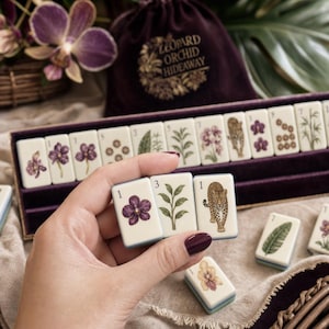 May include: A set of cream-colored mahjong tiles featuring illustrations of purple orchids, green plants, and a leopard. The tiles are displayed in a purple velvet tray alongside a matching drawstring bag labeled Leopard Orchid Hideaway.