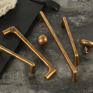 May include: A collection of antique brass cabinet hardware, including handles and a knob. The hardware has a distressed finish and the text "made by Fiona" is printed on each piece. The pieces are arranged on a dark surface.