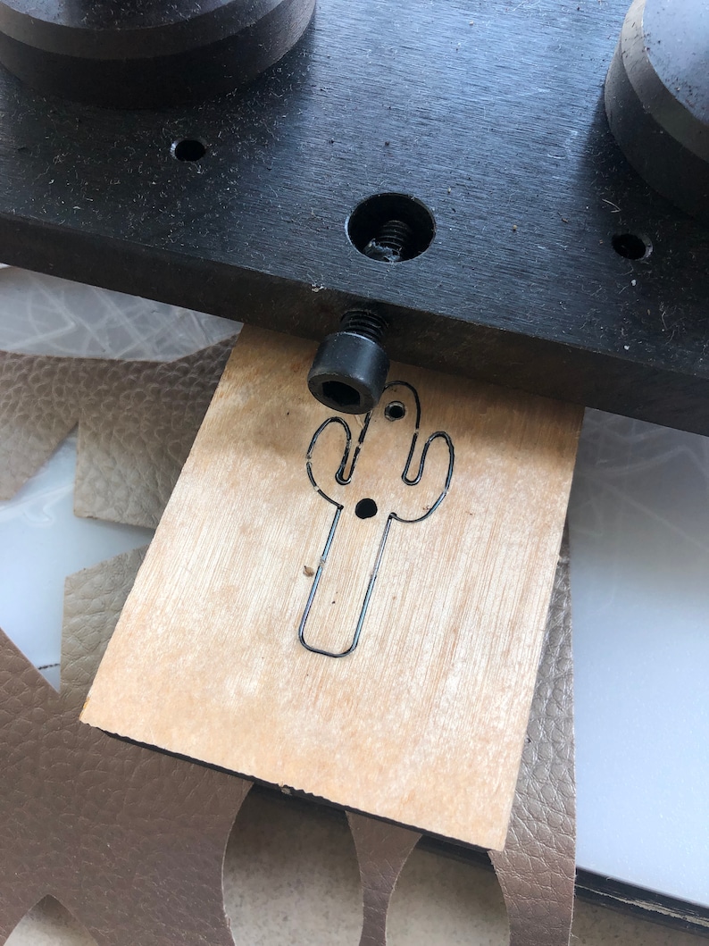 Op de afbeelding: Een close-up van een houten stempel met een cactusontwerp. De stempel wordt gebruikt om een cactusvorm uit een stuk leer te snijden.