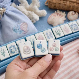 Könnte beinhalten: Ein Set aus weißen Mahjong-Spielsteinen mit blauen Zahlen und Illustrationen von Perlen, Korallen und Meerjungfrauen. Die Spielsteine befinden sich in einem hellblauen Beutel mit Kordelzug und Muschelgrafik, ergänzt durch ein blaues Tablett.