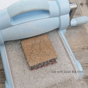 Op de afbeelding: Een lichtblauwe stansmachine met een zilveren, gepatenteerde snijmat. Een bruine stans is op de mat geplaatst. De tekst "Use with Sizzix Big shot" is zichtbaar in de rechterbenedenhoek van de afbeelding.