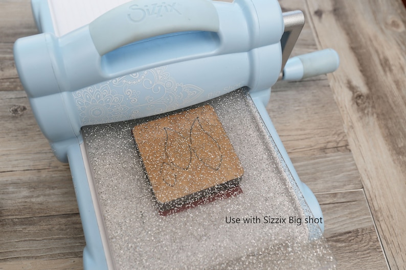 Op de afbeelding: Een lichtblauwe stansmachine met een zilveren, gepatenteerde snijmat. Een bruine stans is op de mat geplaatst. De tekst "Use with Sizzix Big shot" is zichtbaar in de rechterbenedenhoek van de afbeelding.