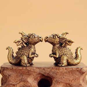 Pode incluir: Duas pequenas figuras de dragão em latão metálico viradas uma para a outra. Cada dragão apresenta escamas detalhadas, pequenas asas, uma cauda curva e olhos grandes. As figuras têm um acabamento texturizado e estão posicionadas lado a lado sobre uma base de madeira.