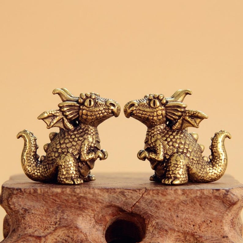 K&ouml;nnte beinhalten: Zwei kleine Drachenfiguren aus Messing, die sich gegen&uuml;berstehen. Jeder Drache hat detaillierte Schuppen, kleine Fl&uuml;gel, einen geschwungenen Schwanz und gro&szlig;e Augen. Die Figuren haben eine strukturierte Oberfl&auml;che und stehen nebeneinander auf einem Holzsockel.