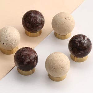 May include: A set of six spherical cabinet knobs featuring a mix of textured beige stone and polished dark brown marble spheres, each mounted on a circular gold-toned metal base. The knobs are arranged in a circular pattern on a flat surface.