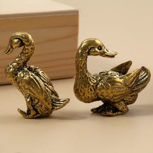 Pode incluir: Duas estatuetas de pato de latão antigo. Um pato está em pé com o pescoço curvado, enquanto o outro está em posição de descanso. Os patos têm texturas de penas detalhadas e estão sobre um fundo neutro.