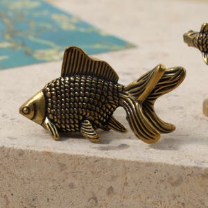 Pode incluir: Dois objetos decorativos antigos em latão em forma de peixe. Um é uma figura de peixe detalhada, e o outro é um selo com um peixe no topo. Os objetos estão em uma superfície de cor clara, com um fundo azul e verde.