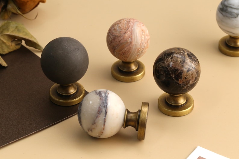Op de afbeelding: Een collectie van vier bolvormige kastknoppen met gepolijste steen- en matte afwerkingen. Elke knop is gemonteerd op een ronde metalen voet in antieke messingkleur. De stenen omvatten een mat antracietgrijs, een gemarmerd roze, een donkerbruin en een wit.