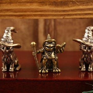 May include: Three bronze figurines: two cats wearing witch hats and a frog wizard holding a staff and book. The figurines are on a dark red surface with a wooden backdrop.