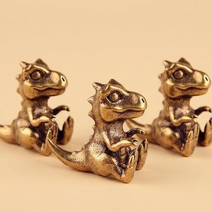 May include: Three small, metallic bronze-coloured dinosaur figurines in a seated position. Each dinosaur features a textured surface, a prominent tail, short arms, and a detailed head with large eyes, arranged in a triangular formation.