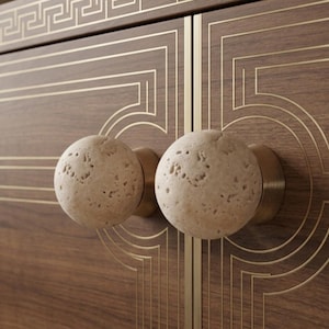 May include: Two spherical, porous, beige stone cabinet knobs mounted on a dark brown wood cabinet door. The cabinet features thin, inlaid gold-colored metal lines forming geometric patterns and a Greek key border along the top edge.
