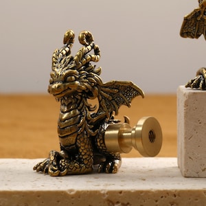 Op de afbeelding: Gedetailleerd messing drakenbeeldje met een knop bevestigd, mogelijk voor een kast of lade. De draak zit in een zittende positie met vleugels en ingewikkelde schubben. De afwerking is een warme, antieke goudkleur.