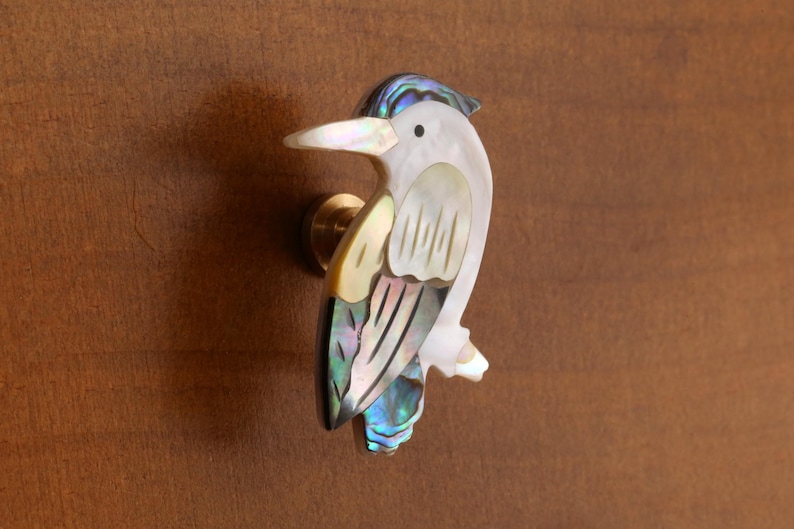 May include: A decorative bird-shaped cabinet knob. The knob features a white body with iridescent shell accents in blue, green, and gold. The bird has a white beak and a brass mounting base. The knob is mounted on a brown surface.