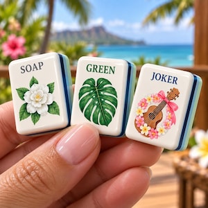 Pode incluir: Três peças retangulares de mahjong com faces brancas e lados azulados. A primeira peça apresenta uma flor branca e o texto SOAP. A segunda mostra uma folha de monstera verde e o texto GREEN. A terceira mostra um ukulele e o texto JOKER.